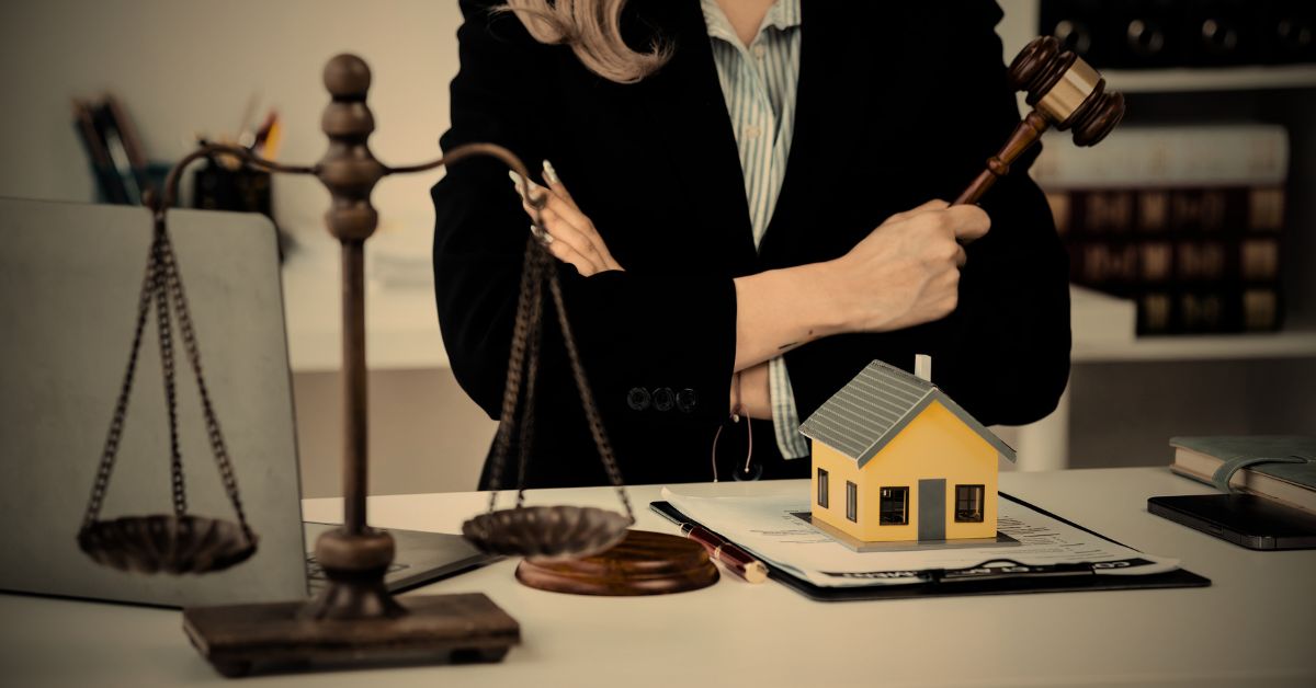Attorney holding a gavel standing behind a desk with a house model and legal documents.