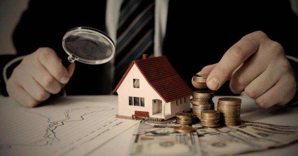 Person reviewing home insurance documents with a model house and stacked coins on a desk.