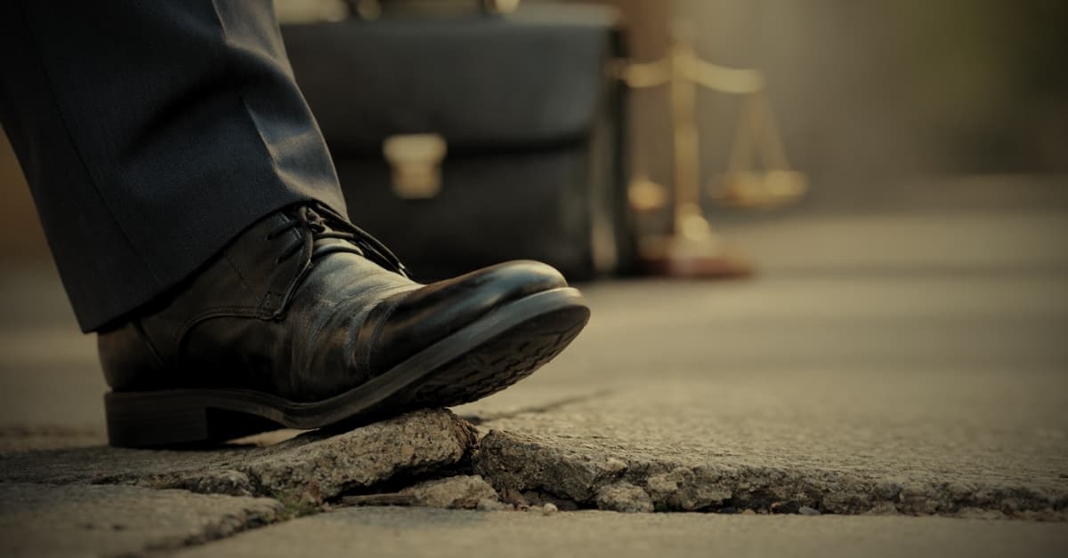 Business shoe stepping on a cracked and uneven sidewalk creating a trip hazard